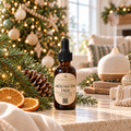 An amber glass dropper bottle labeled 'Around the Tree' by Chandelle & Savon, styled on a marble surface with Christmas greenery, pinecones, dried orange slices, and a diffuser in the background.