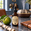 A bottle of Chandelle & Savon Bergamot Leather essential oil set on a dark stone tabletop, surrounded by a fresh bergamot citrus, a birch branch, a brown leather folio, and a wooden diffuser in the background.