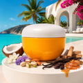 A round cosmetic jar with a white lid and a bright yellow-orange body sits on a white table in a tropical beach setting. The jar is styled with coconut, almonds, vanilla beans, wood chips, and honey, with turquoise water, palm trees, and a white resort building in the background.