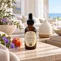 An amber glass dropper bottle labeled 'CHANDELLE & SAVON COASTAL' sits on a stone surface in a bright, styled interior. A white diffuser, flowers, a peach, and a sofa are visible in the background, suggesting a home fragrance or wellness product scene.