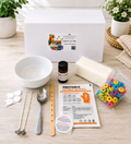 A DIY candle making kit laid out on a light wood surface with a white branded box behind it. Visible items include a white bowl, metal wicks, a spoon, wooden wick holder, printed instruction card, a small bottle of citrus oil, a bar of soap, a bag of colorful ring-shaped candies, and a pair of nitrile gloves.