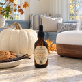 An amber glass dropper bottle of Chandelle & Savon Pumpkin Cherie diffuser oil stands on a stone countertop in a cozy autumn kitchen. A large white pumpkin, cookies, vanilla beans, and a wooden diffuser are arranged around it, with a blurred living room background featuring pillows and a window.