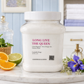 A large white jar of Chandelle & Savon laundry soap labeled 'LONG LIVE THE QUEEN' sits on a marble surface in a bright, styled bathroom setting with citrus slices, flowers, and folded towels in the background.