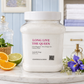 A large white jar of Chandelle & Savon laundry soap labeled 'LONG LIVE THE QUEEN' sits on a marble surface in a bright, styled bathroom setting with citrus slices, flowers, and folded towels in the background.