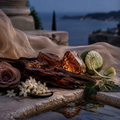 Amber resin crystals and a dark wood slice arranged on a stone surface with a draped sheer fabric, a pink rose, white flowers, mint leaves, and a lime slice, set against an outdoor waterfront at dusk.