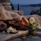 Amber resin crystals and a dark wood slice arranged on a stone surface with a draped sheer fabric, a pink rose, white flowers, mint leaves, and a lime slice, set against an outdoor waterfront at dusk.