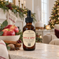 An amber glass dropper bottle of Chandelle & Savon Apple Spice massage oil stands on a marble surface in a cozy holiday living room. The bottle has a black dropper cap and a cream label with red text.