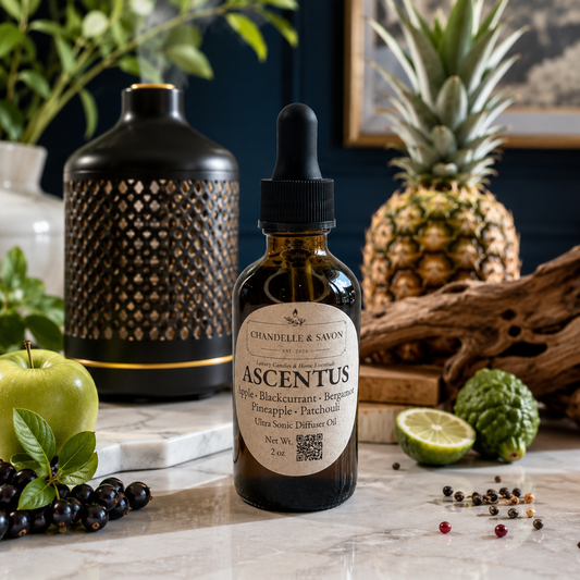 A bottle of Chandelier & Savon Ascentus citrus oil stands on a marble surface surrounded by fruit and decorative props, including a pineapple, green apple, blackcurrants, a patterned black and gold diffuser, and driftwood.