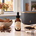 An amber glass dropper bottle labeled 'Chandelle & Savon' and 'Autumn Delight' sits on a kitchen counter beside a gray diffuser, cinnamon sticks, cloves, and a spoonful of liquid.