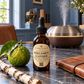 A bottle of Chandelle & Savon Bergamot Leather essential oil set on a dark stone tabletop, surrounded by a fresh bergamot citrus, a birch branch, a brown leather folio, and a wooden diffuser in the background.
