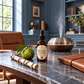 A bottle of Bergamot Leather oil by The Perfume Shop sits on a marble-topped desk in a styled home office. The amber dropper bottle is surrounded by a green bergamot fruit, a piece of birch wood, and a dark brown leather portfolio notebook.