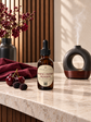 A bottle of Chandelle & Savon Black Cherry Reserve diffuser oil stands on a marble countertop, styled with fresh cherries and blackberries, a dark red cloth, a diffuser, and a vase of flowers in a warm indoor setting.