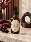Chanelle & Savon Black Cherry Reserve diffuser oil in an amber glass dropper bottle, styled on a marble countertop with fresh cherries, blackberries, a dark red cloth, and a diffuser in the background.