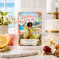 A wax melt package labeled 'Brazilian Creme' by Chandelle & Savon, displayed in a clear plastic clamshell on a marble surface with tropical beach artwork on the front. The scene includes a lemon half, pink roses, a cherry, and a softly blurred coastal interior background.