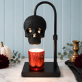 A black skull-shaped hanging lamp with a rectangular base, shown above a glowing red glittery candle holder on a marble surface. The scene is styled with a white pumpkin, flowers, greenery, and a light blue paneled wall in the background.