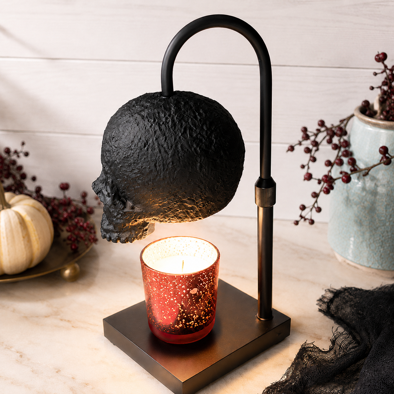 A black skull-shaped Candle Warmer hovers over a red glitter candle holder on a square black base, styled on a marble surface with pumpkins, dried berries, and a ceramic vase in the background.