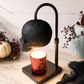 A black skull-shaped Candle Warmer hovers over a red glitter candle holder on a square black base, styled on a marble surface with pumpkins, dried berries, and a ceramic vase in the background.