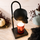 A Halloween-themed skull-shaped table lamp with a black metal frame and a textured black skull shade, holding a lit red glitter candle. The lamp is styled on a light wood surface with seasonal props including a small pumpkin, dried berries, and a ceramic vase.