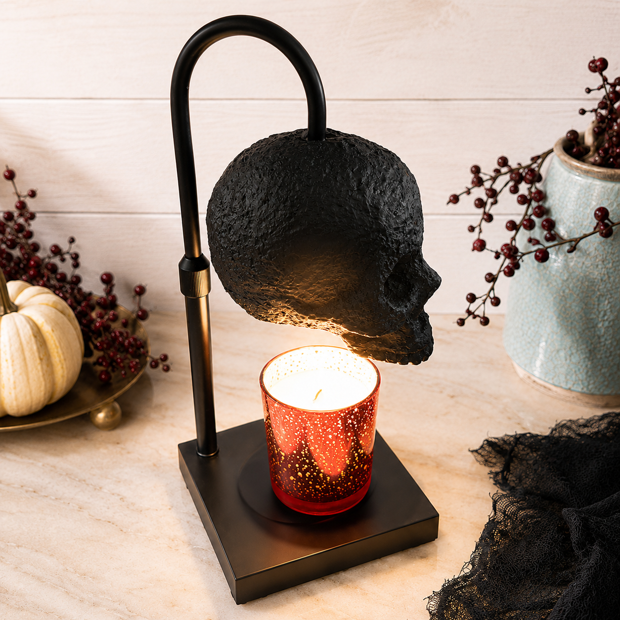 A Halloween-themed skull-shaped table lamp with a black metal frame and a textured black skull shade, holding a lit red glitter candle. The lamp is styled on a light wood surface with seasonal props including a small pumpkin, dried berries, and a ceramic vase.