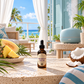 A bottle of Caribbean Crush diffuser oil dropper sits on a speckled stone table in a tropical beach patio setting. The scene includes a light blue diffuser, a sliced melon, pineapple, a halved coconut, and a blue-and-white striped towel, with a bright ocean and palm trees in the background.
