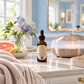 A bottle of Cashmere Petals diffuser oil by Chandelle & Savon stands on a marble bathroom counter next to a glowing diffuser, with a vase of flowers and a folded cream knit blanket nearby.