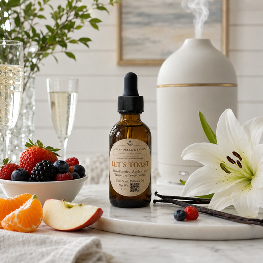An amber glass dropper bottle labeled 'Chandelle & Savon' and 'Let's Toast' fragrance oil, styled on a marble tray with strawberries, blueberries, orange slices, an apple, vanilla beans, a white lily flower, a diffuser, and champagne glasses in a bright kitchen setting.