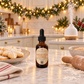 An amber glass dropper bottle labeled 'Chandelle & Savons Christmas Cookie' stands on a kitchen counter surrounded by holiday baking props, including a plate of decorated cookies, a rolling pin, flour, and a diffuser in the background.