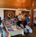 A rustic indoor event space with wooden ceiling beams, hanging lights, and multiple dining tables set with tablecloths and glassware. Several people are standing and sitting around the tables, and wall decor including framed art and a large star motif is visible.