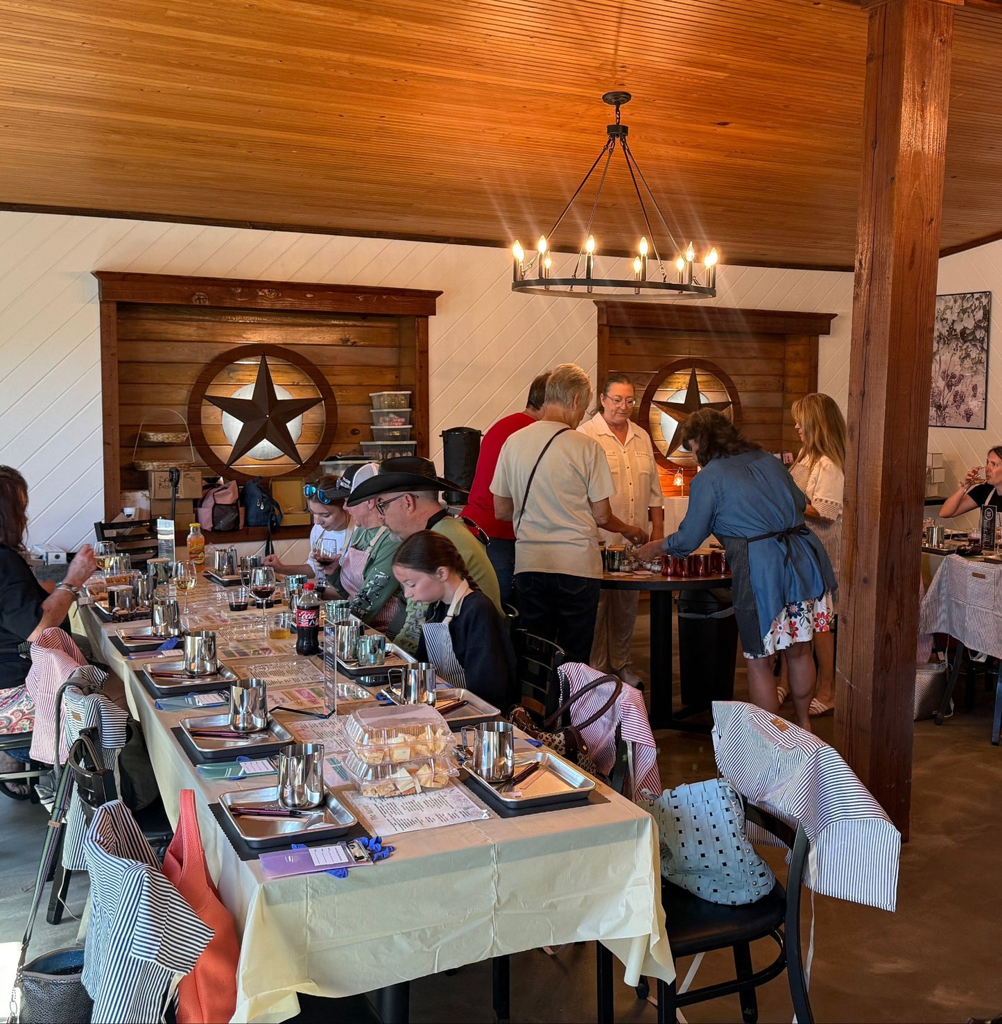 A rustic indoor event space with wooden ceiling beams, hanging lights, and multiple dining tables set with tablecloths and glassware. Several people are standing and sitting around the tables, and wall decor including framed art and a large star motif is visible.