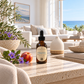 A bottle of Coastal diffuser oil by Chandelle & Savon sits on a stone coffee table in a bright, modern living room. The amber glass bottle has a black dropper cap and a cream label.