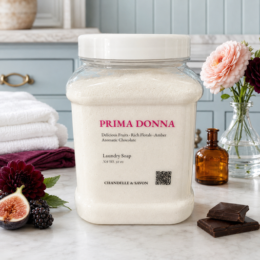 A large clear plastic jar of Prima Donna Laundry Soap filled with white soap powder, set on a marble countertop with folded towels, flowers, and chocolate pieces in a kitchen-like setting.