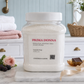 A large clear plastic jar of Prima Donna Laundry Soap filled with white soap flakes, set on a marble board in a styled bathroom setting with folded towels, a pink rose, and a chocolate bar nearby.
