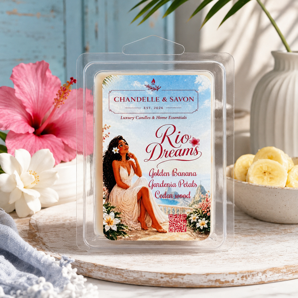 A packaged wax melt or scented melt product labeled 'Chandelle & Savon Rio Dreams' sits on a rustic wooden surface with tropical props including a pink hibiscus flower, white flowers, banana slices, and a vase in the background.