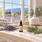 A bottle of Fields of Lavandender diffuser oil sits on a speckled beige countertop in a bright kitchen or spa setting. The scene is styled with lavender sprigs, rosemary, a white ceramic diffuser, and a striped towel, with a large window behind showing a scenic lavender field and mountains.