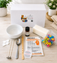 A DIY candle making kit laid out on a light wood surface with a white branded box behind it. Visible items include a white bowl, metal wicks, a spoon, wooden wick holder, printed instruction card, a small bottle of citrus oil, a bar of soap, a bag of colorful ring-shaped candies, and a pair of nitrile gloves.