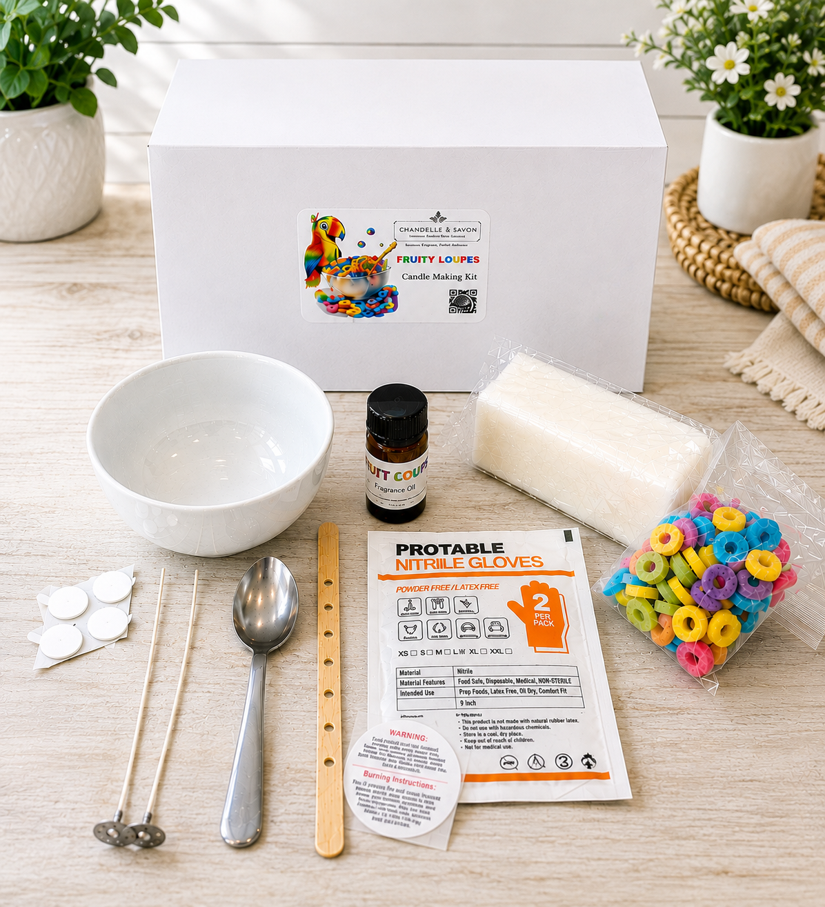 A DIY candle making kit laid out on a light wood surface with a white branded box behind it. Visible items include a white bowl, metal wicks, a spoon, wooden wick holder, printed instruction card, a small bottle of citrus oil, a bar of soap, a bag of colorful ring-shaped candies, and a pair of nitrile gloves.