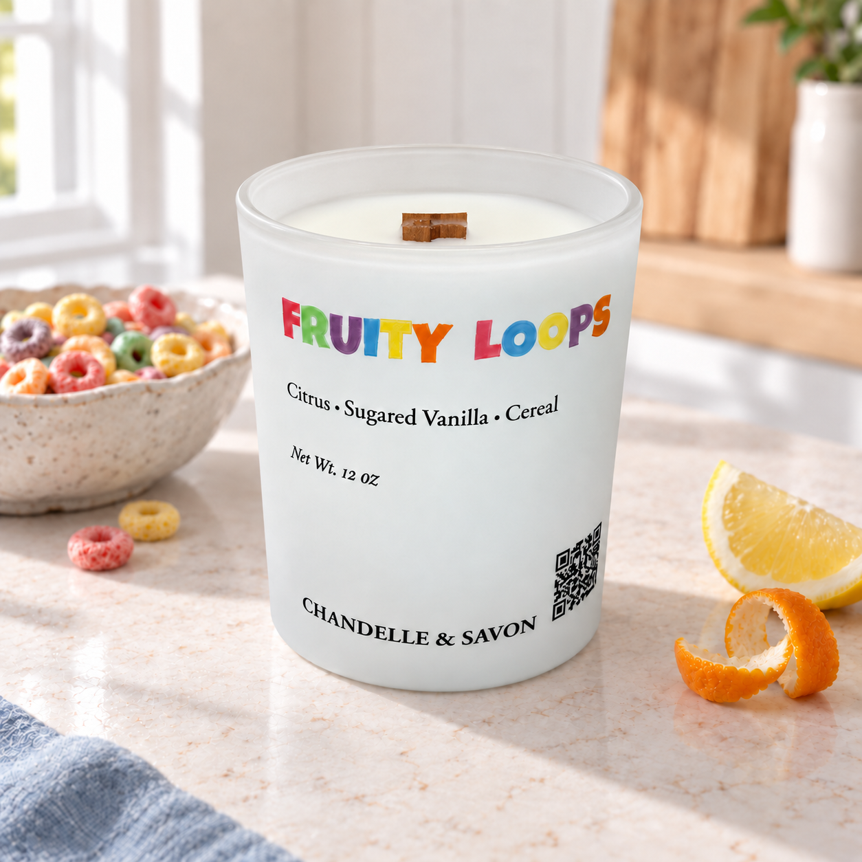 A frosted glass candle jar labeled 'FRUITY LOOPS' with a small wooden wick, set on a kitchen counter with cereal, a lemon slice, and an orange peel nearby. The candle is branded CHANDELL & SAVON.