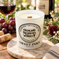 A white glass candle jar labeled Highland Reserves Winery Sweet Inks sits on a marble board outdoors on a wooden table, surrounded by grapes, a glass of red wine, flowers, and a wine bottle.