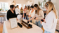 A group of women gather around a table displaying a candle bar setup with multiple small bottles and black candle holders in a bright, modern indoor space.