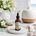 A small amber glass dropper bottle labeled Honey Silk by Chandelle & Savon sits on a white marble tray. The scene is styled with white flowers in a vase, driftwood, a beige cloth, and a white diffuser in the background.
