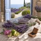 Lavender bundles, a bowl of lavender, pink flowers, lime slices, and a small wooden box of dark stones arranged on a rustic table with a linen cloth, set in front of an open blue door overlooking a lavender field and the sea.
