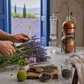 A person's hands are shown harvesting fresh lavender with scissors at a rustic table. On the table are a copper distillation apparatus, a glass jar, a small bowl of dried lavender buds, a lime, and a wooden cutting board.