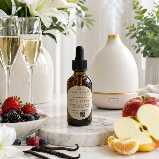 An amber glass dropper bottle labeled 'Chandelle & Savon' and 'Let's Toast' sits on a marble slab in a styled kitchen scene with a white diffuser, champagne flutes, fruit, and flowers.