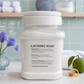 A clear plastic jar of Chandelle & Savon Laundry Soap filled with white soap flakes, set on a marble countertop with a green lime, a purple flower, and a folded towel in the background.