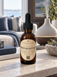 An amber glass dropper bottle labeled 'Chandelle & Savon Midnight Ice' sits on a light stone countertop in a softly blurred living room setting with a sofa, diffuser, and window in the background.