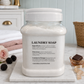 A large clear plastic jar of Chandeliers & Savon laundry soap with a white screw-top lid, displayed on a white marble board. The jar is set on a marble surface with a pink rose, blackberries, and a small dish of soap in a softly lit bedroom setting.