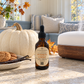 An amber glass dropper bottle of Chandelle & Savon Pumpkin Cherie diffuser oil stands on a stone countertop in a cozy autumn kitchen. A large white pumpkin, cookies, vanilla beans, and a wooden diffuser are arranged around it, with a blurred living room background featuring pillows and a window.