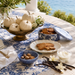 An outdoor dining table styled with a blue and white patterned tablecloth, white pumpkins in a bronze bowl, cookies on plates, vanilla beans, cream, and a blue floral cookie tin with its lid leaning beside it. A real environment with greenery and water is visible in the background.