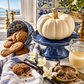 A styled autumn kitchen scene with a white decorative pumpkin on a blue pedestal, blue-and-white ceramic cookies, brown cookies spilling out, vanilla beans, white flowers, a glass of milk, and a brass dish on a marble countertop near a window.
