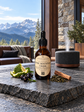 An amber glass dropper bottle labeled 'Steel Mountain Creek' by 'Chandelle & Savon' sits on a dark stone slab in a cozy indoor setting with a mountain landscape visible through large windows. A diffuser, cinnamon stick, dried herbs, and citrus slices are arranged around it.